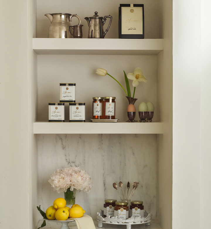 White shelf with items including jars of honey and fruit spread, packages of tea, lemons, a silver pitcher, and flowers.