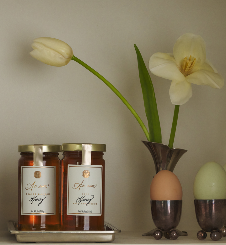 Two jars of honey on a tray with a vase containing flowers and two eggs in egg cups against a plain wall.