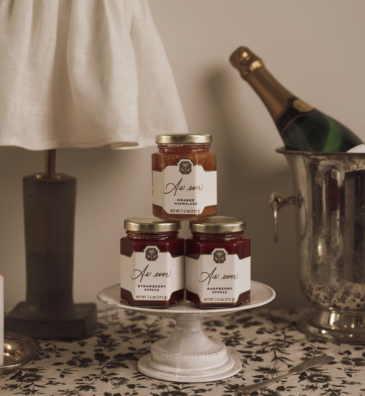 Three jars of fruit spread stacked on a ceramic plate beside an unlit lamp, and a bottle of champagne in a silver ice bucket. 