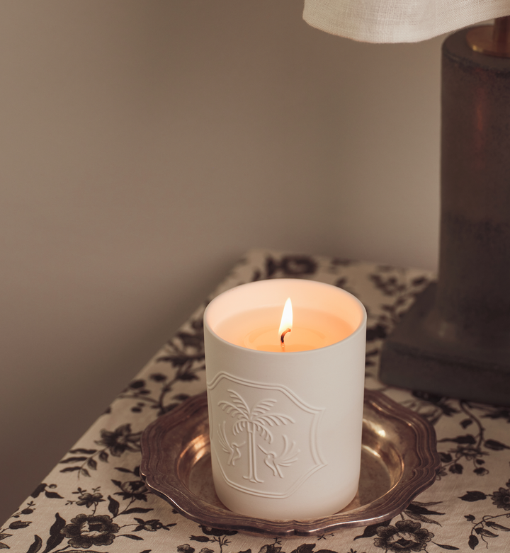 Floral surface with a white lit candle on a small silver tray and a lamp. 