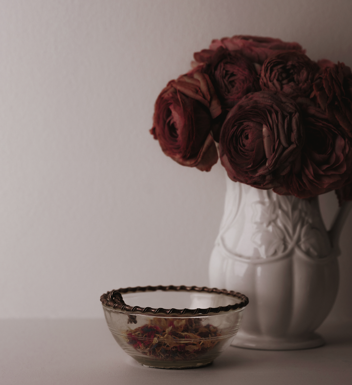 White vase with red flowers and a small glass bowl filled with petals on a light surface.