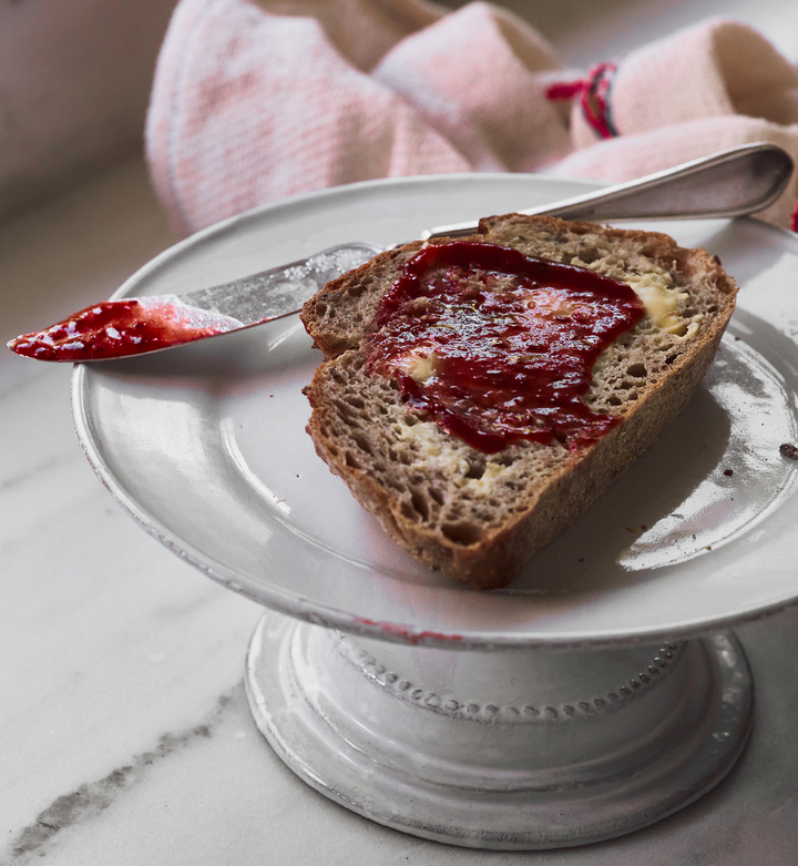 Slice of bread with red fruit spread on a white plate with a knife, on a marble surface.