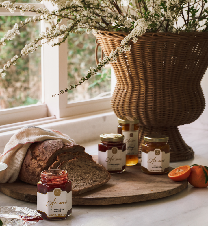 A marble countertop with sliced bread on a wooden board, jars of honey and fruit spread, an orange, and flowers.