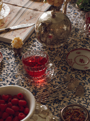 Clear teacup with red tea, silver teapot, and bowl of raspberries, on a patterned tablecloth.