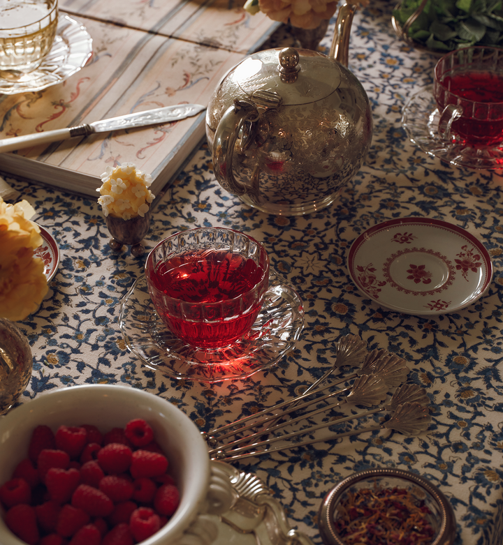 Clear teacup with red tea, silver teapot, and bowl of raspberries, on a patterned tablecloth.