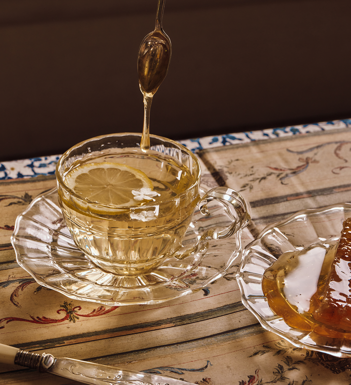 Honey being drizzled into a glass teacup with a lemon slice on a decorative table.