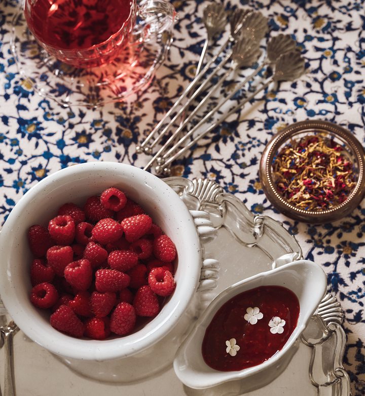 Bowl of raspberries and fruit spread in a ceramic bowl on a silver tray beside a glass bowl of flower sprinkles on a patterned tablecloth.