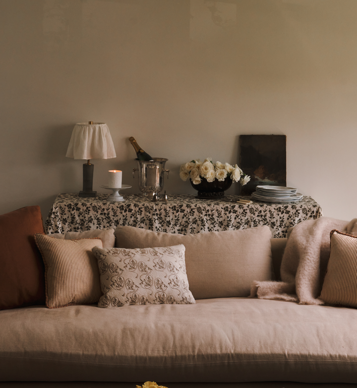 A sideboard covered in a floral table cloth with a lamp, candle, champagne in a silver ice bucket, white flowers, and a stack of plates. A beige sofa with decorative pillows is in front. 