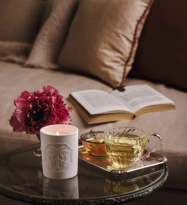 Candle, flower, and teacup on a glass table with an open book on a blurred sofa in the background