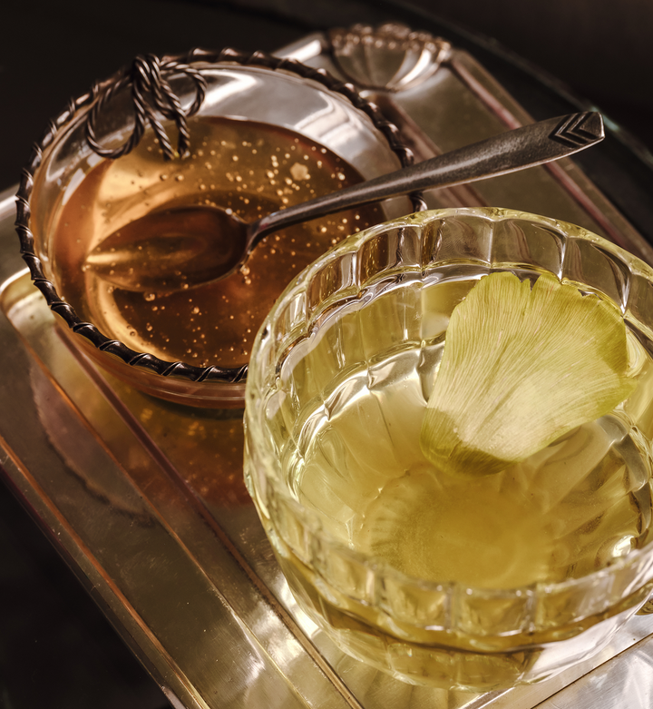 Glass of yellow liquid with a leaf in it, beside a clear bowl of honey on a silver tray