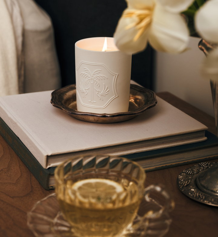 Candle with decorative emblem on a book, accompanied by a glass of tea with lemon, and flowers.