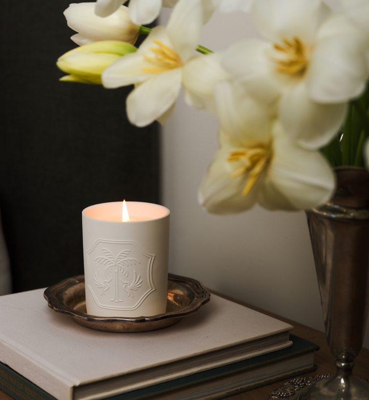 White candle with decorative emblem on a stack of books, beside white flowers.