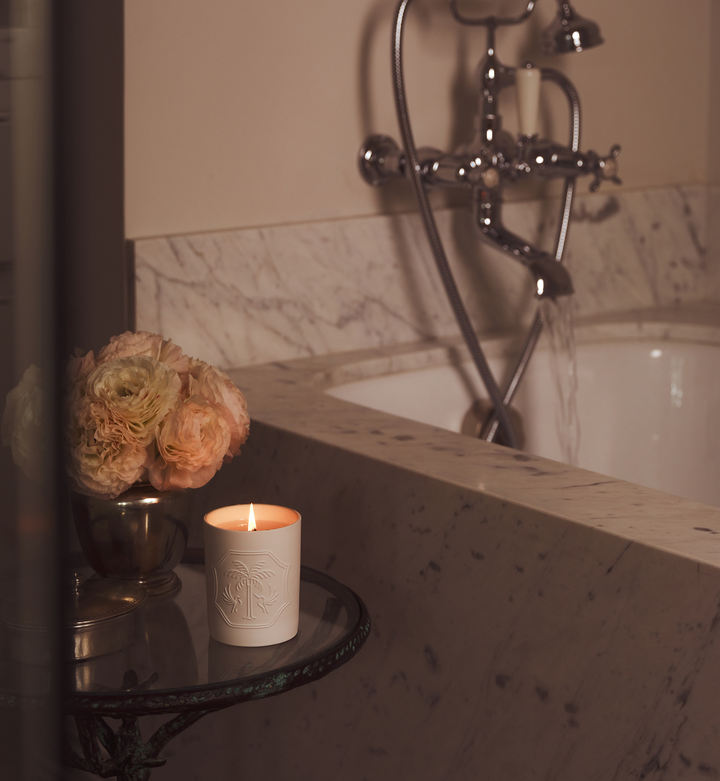 Candle and flowers on a glass and iron table beside a marble bathtub. 