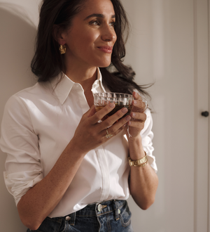 Smiling woman holding a glass of tea, leaning against a white wall. 
