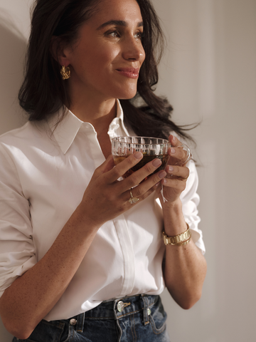 Smiling woman holding a glass of tea, leaning against a white wall. 