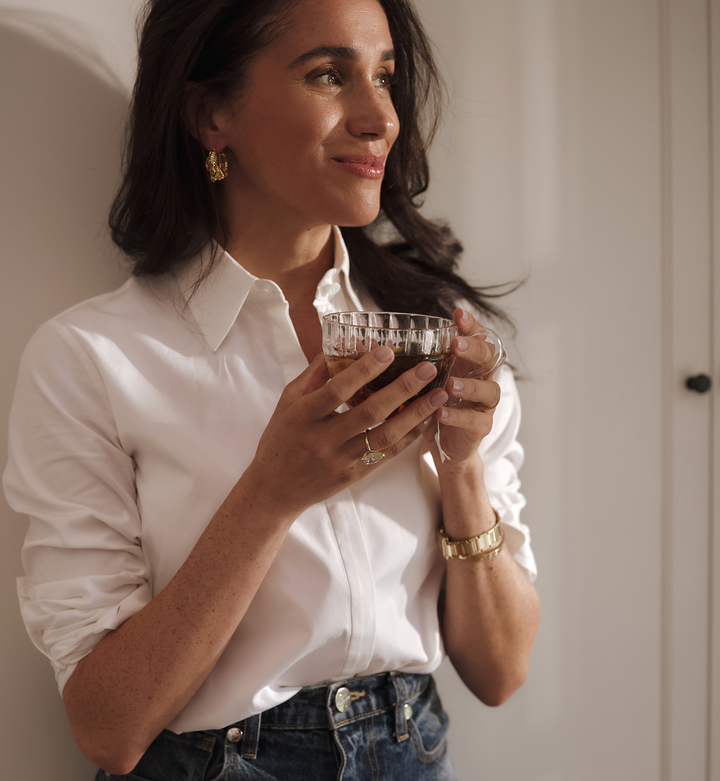 Smiling woman holding a glass of tea, leaning against a white wall. 