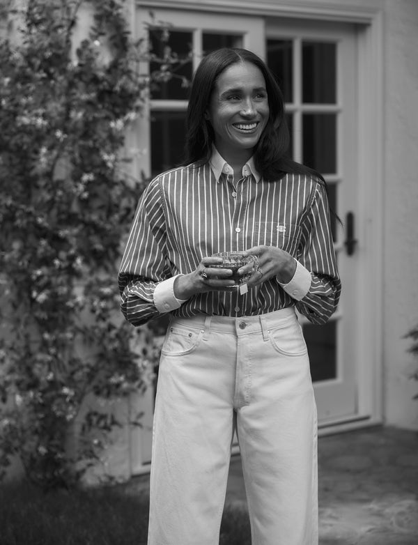 A woman standing in front of a house, smiling, holding a cup of tea in a glass mug. 