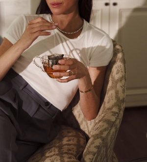 Woman sitting on a patterned beige chair dipping a tea bag into a clear cup of tea. 