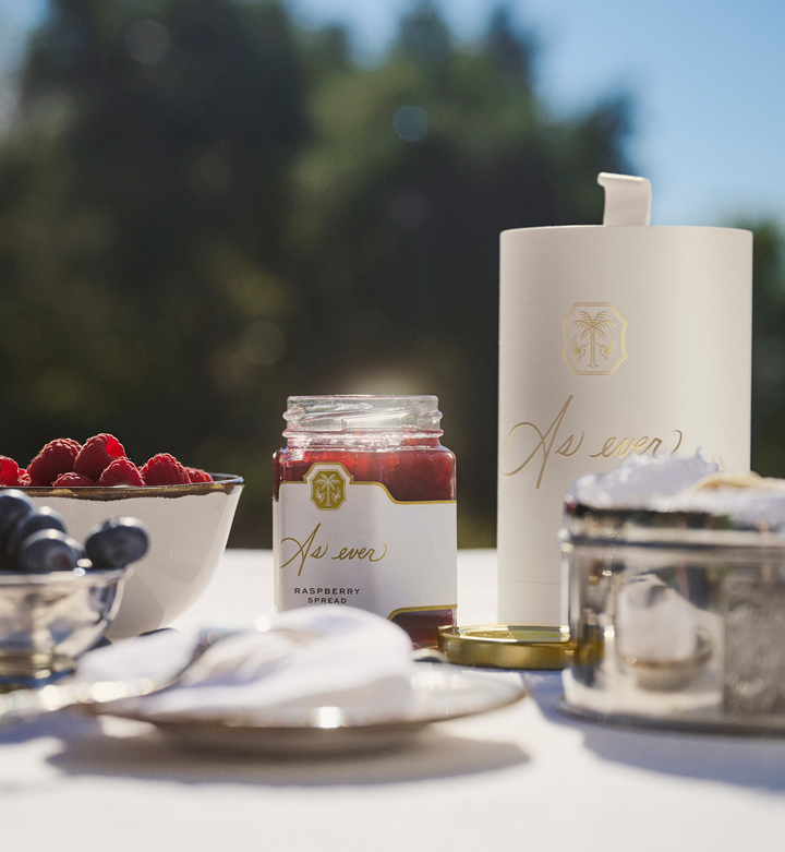 Outdoor table setting with a jar of raspberry spread, a beige cylindrical box, and bowls of berries.