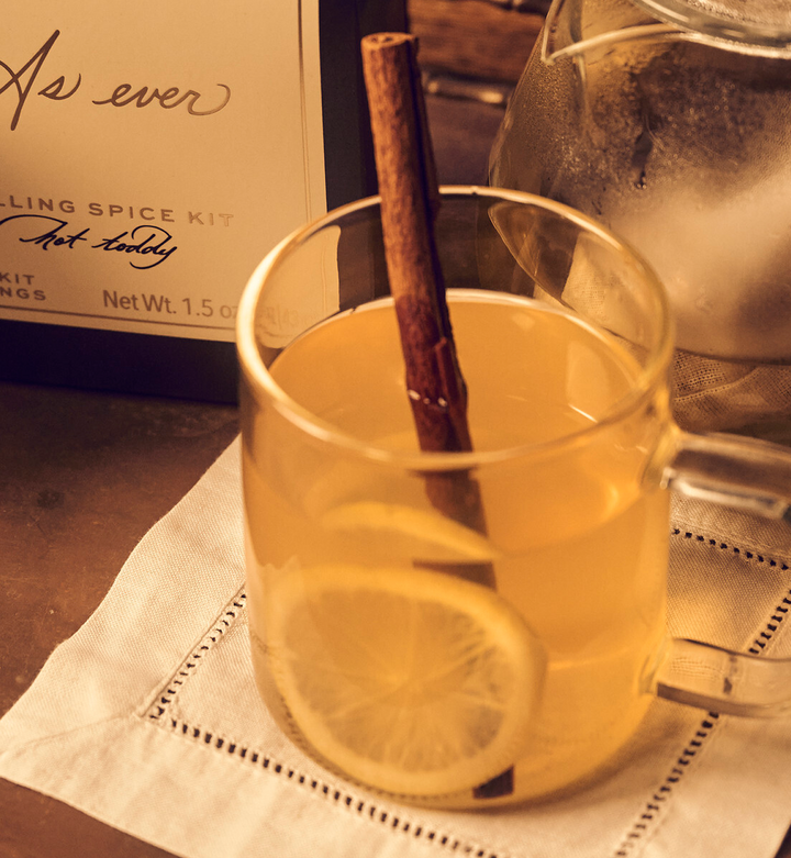 Glass of hot toddy with lemon slices and a cinnamon stick, next to a spice kit.