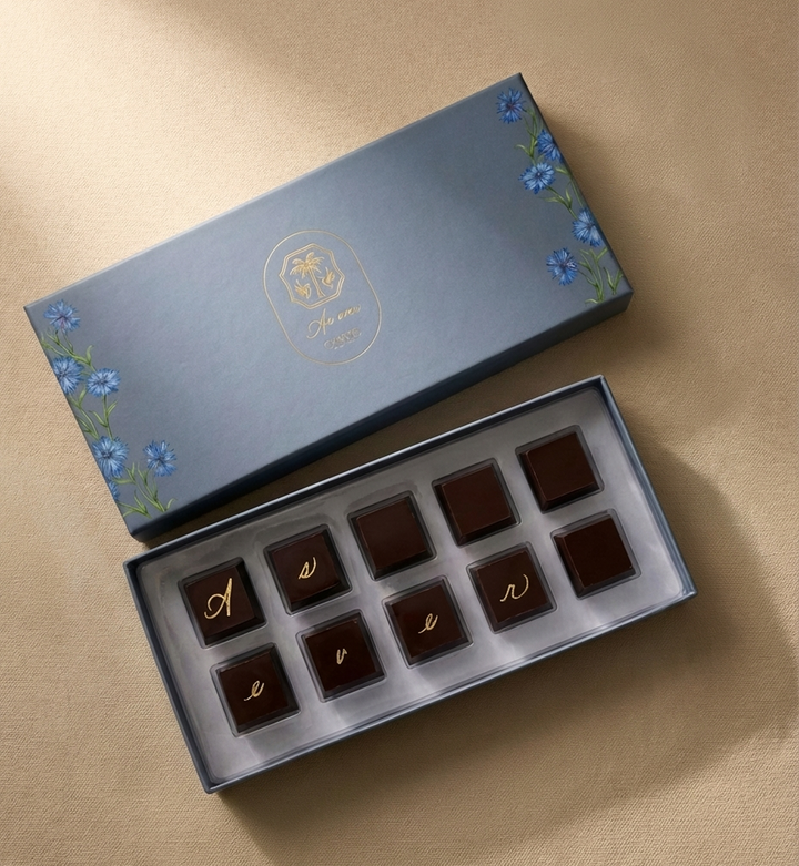 An open box of chocolates on which the lid has blue flowers on either edge and a logo in gold on the centre. Inside the box are 10 dark chocolates, six of which have letters on them spelling out As ever. 