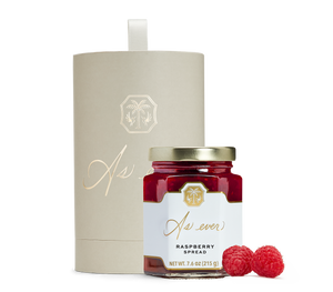 A jar of raspberry spread, raspberries, and a beige cylindrical box with gold detailing.