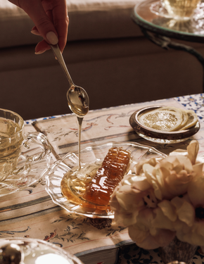 Honey being drizzled from a spoon into a glass dish on a table with a teacup and flowers.