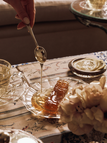 Honey being drizzled from a spoon into a glass dish on a table with a teacup and flowers.