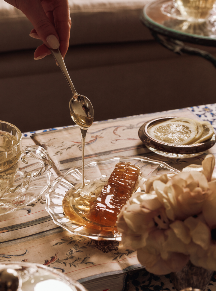 Honey being drizzled from a spoon into a glass dish on a table with a teacup and flowers.