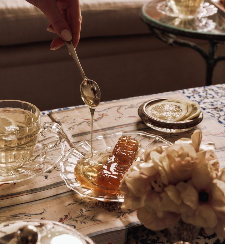 Honey being drizzled from a spoon into a glass dish on a table with a teacup and flowers.