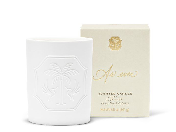 An unlit white candle with an engraved design beside a beige and gold candle box.