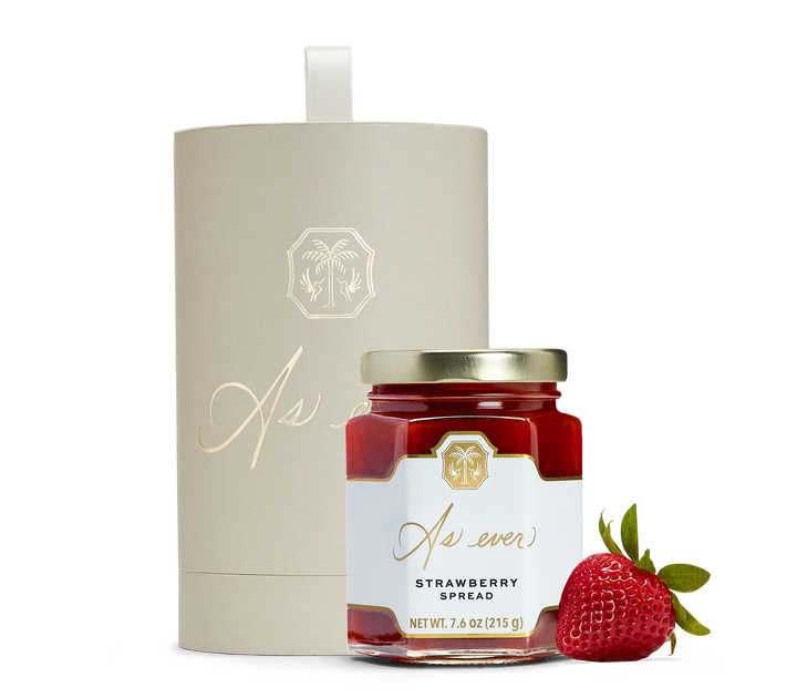 A jar of strawberry spread, a strawberry, and a beige cylindrical box with gold detailing. 