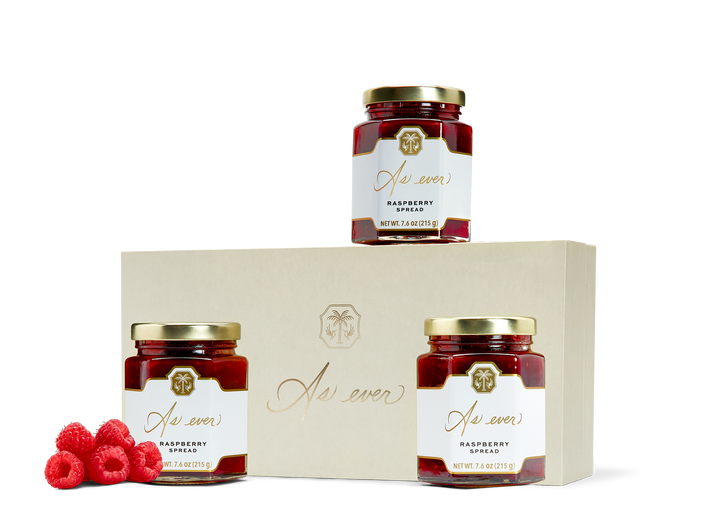 Three jars of raspberry spread, loose raspberries, and a beige box with a gold logo. 
