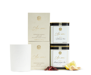 An unlit white candle beside two beige and gold candle boxes, two cans of As ever tea, and loose lemon, ginger, and hibiscus. 