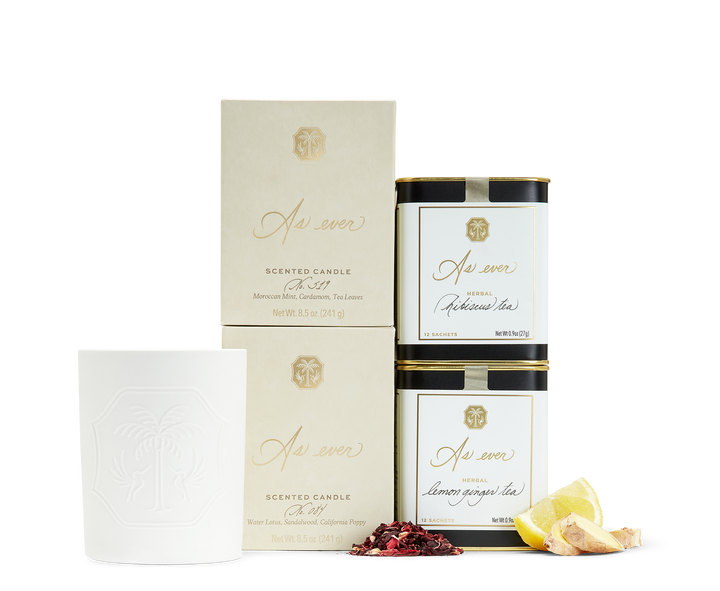 An unlit white candle beside two beige and gold candle boxes, two cans of As ever tea, and loose lemon, ginger, and hibiscus. 