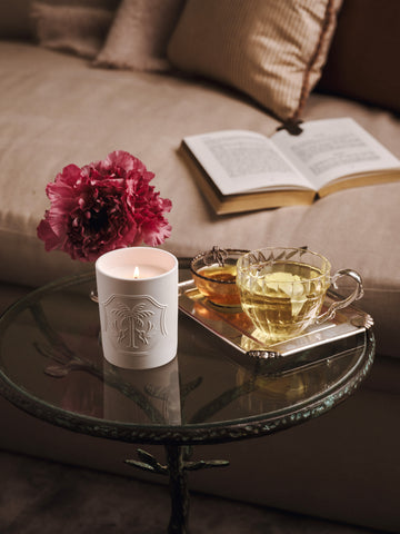 A lit white candle, a pink flower, and silver tray with tea, on a glass table in front of a couch with an open book on it. 