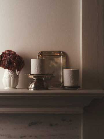 Candles, a vase, and a silver tray on a mantelpiece against a plain wall.