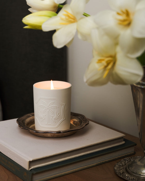 White candle with decorative emblem on a stack of books, beside white flowers.