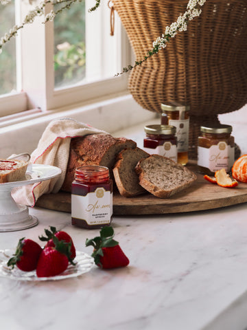 A marble countertop with sliced bread on a wooden board, jars of honey and fruit spread, and fresh fruit. 