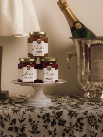Three jars of fruit spread stacked on a ceramic dish beside a bottle of champagne in a silver ice bucket on a floral tablecloth. 