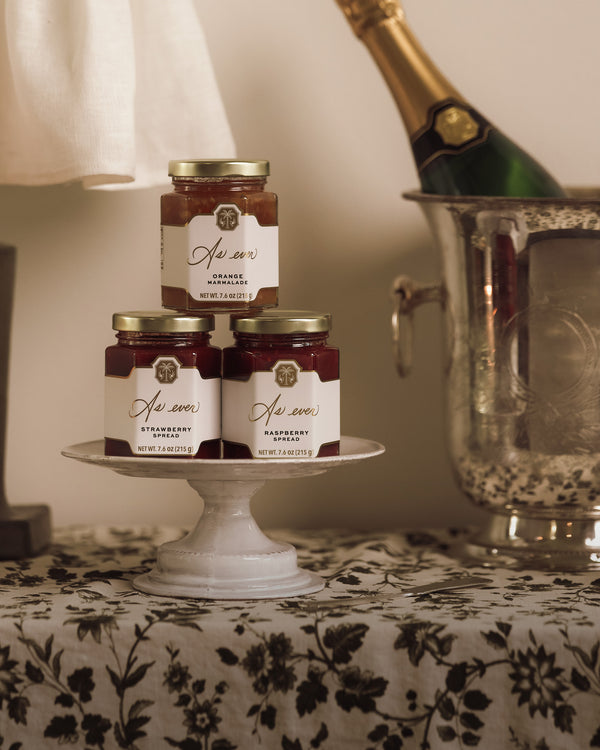 Three jars of fruit spread stacked on a ceramic dish beside a bottle of champagne in a silver ice bucket on a floral tablecloth. 