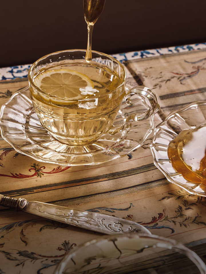 honey being dripped into Tea in a glass teacup with lemon slices on a decorative table.