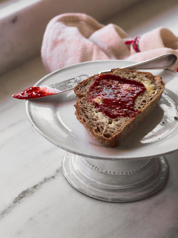 Slice of bread with red fruit spread on a white plate with a pink cloth in the background.