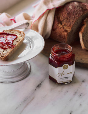 A jar of raspberry spread on a table with bread. 