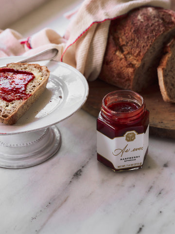 A jar of raspberry spread on a table with bread. 
